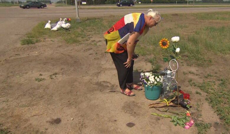 A woman wearing a colourful shirt, dark slacks and sandles is bending over, her hands on her knees, scanning a small memorial with various flowers. The memorial is set up at an intersection next to a highway.