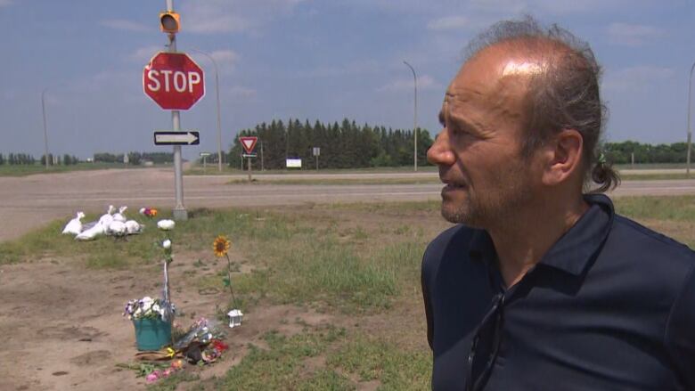 A man with thinning is wearing a navy blue polo shirt. He is standing a few feet away from a small grouping of flowers that have been laid on the dirt, beside a highway intersection. A stop sign and one way sign stand at the corner of the intersection.