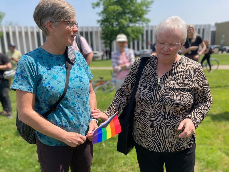 Woman with eyes closed, rests hand on the arm of another woman holding a small rainbow flag