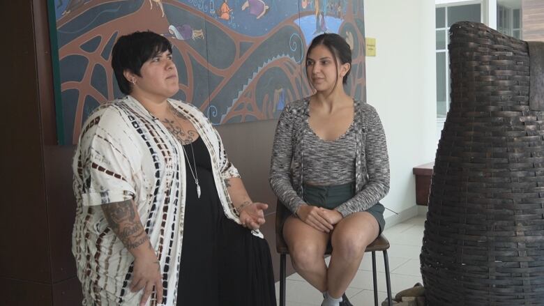 Two women sit in front of painting and are talking.