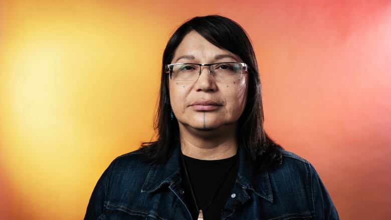 A woman with long black hair, wearing glasses and a black shirt and denim jacket, stares into the camera.