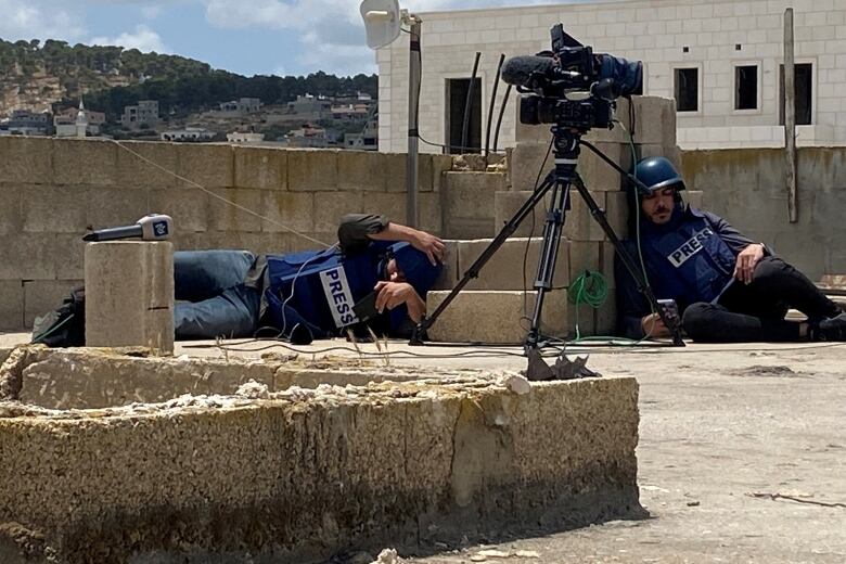 Journalists wearing helmets and bulletproof vests take cover near brick structures as a camera on a tripod and microphone as seen discarded.