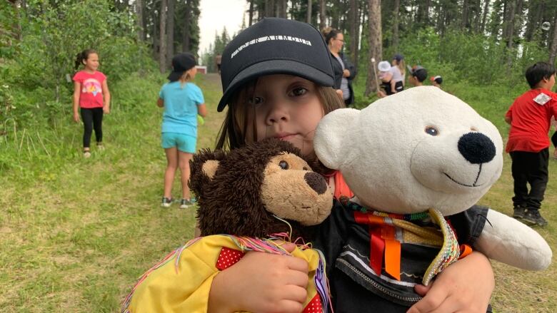 A little girl in a ball cap holds two teddy bears wearing Indigenous regalia.