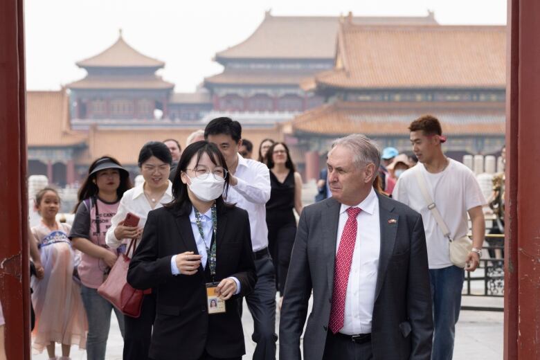 A man wearing a grey suit and red tie walks through a red metal arch, as a group of people follow behind. In the background are a small cluster of ornately decorated buildings with terracotta roofs.