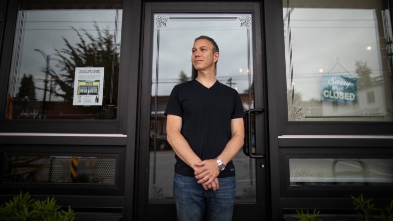 A man in a black T-shirt and jeans stands outside a restaurant looking away from the camera.