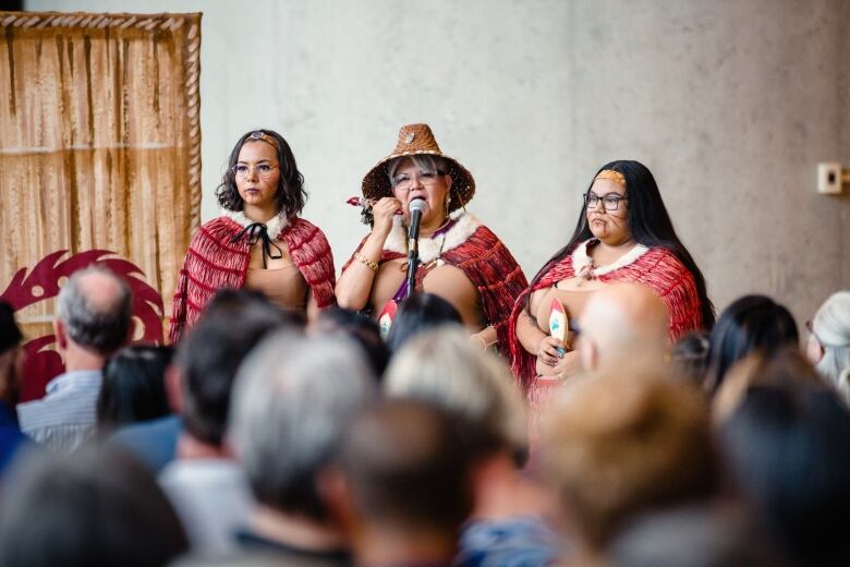 Three Indigenous women in traditional attire addressing a large crowd. 