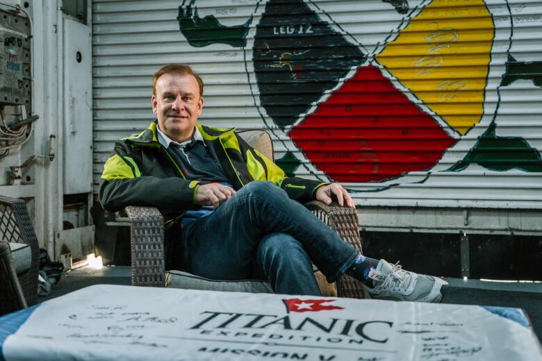 A man wearing a black and yellow jacket sitting in a wicker arm chair. There's a flag in front of him that says Titanic Expedition.