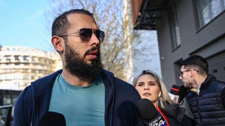 A bearded man in sunglasses wearing a hoodie is shown in an outdoor photo, surrounded by microphones.