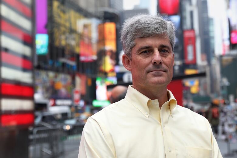 A man wearing a yellowish button-up shirt. He has a clean-shaven face and medium length grey hair.