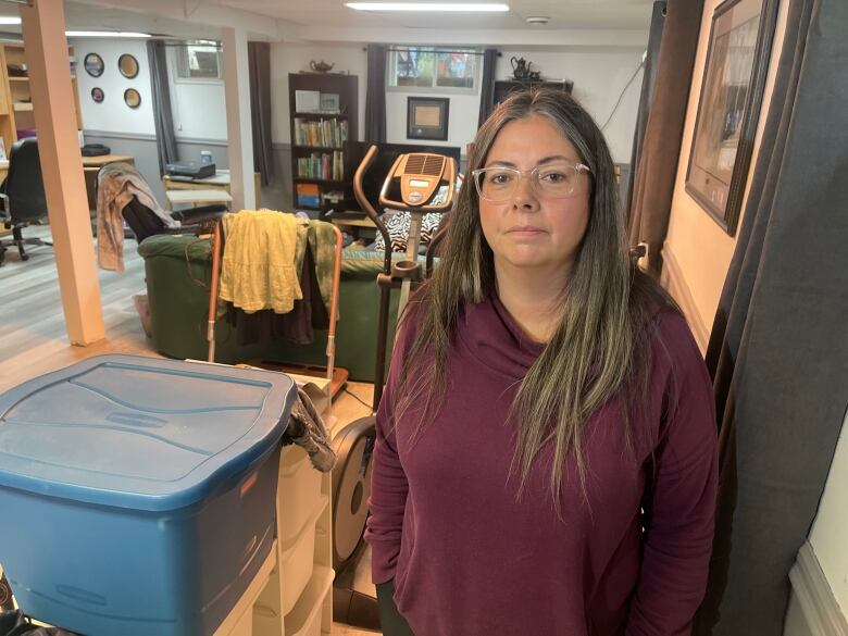 A woman with long brown hair in a purple hoodie stands in a basement filled with boxes. 