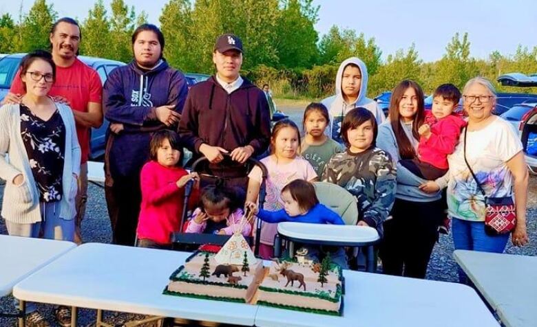 A large Cree family of 13 stands smiling at the camera with a large cake in front of them.