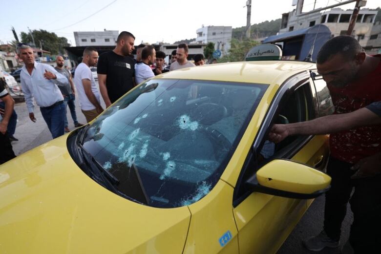 A yellow car with bullet holes in the windshield is inspected. 