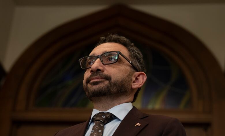 A man wearing glasses and a dark brown suit and tie looks forward, while standing in front of an ornate wooden wall.