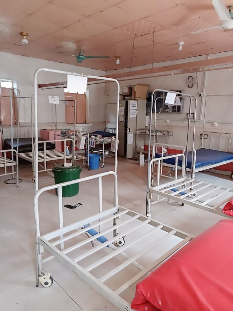 There are several hospital beds in the beige room. Most appear stripped down. There is an open locker in the back, and medical carts in the corner. 