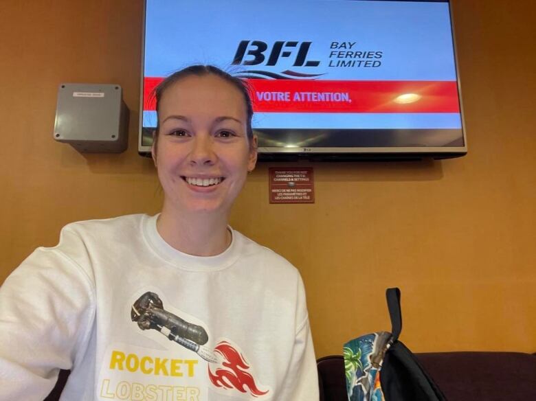 A woman smiles at the camera. There's a television screen behind her. She wears a white sweather that says, 'Rocket Lobster.'