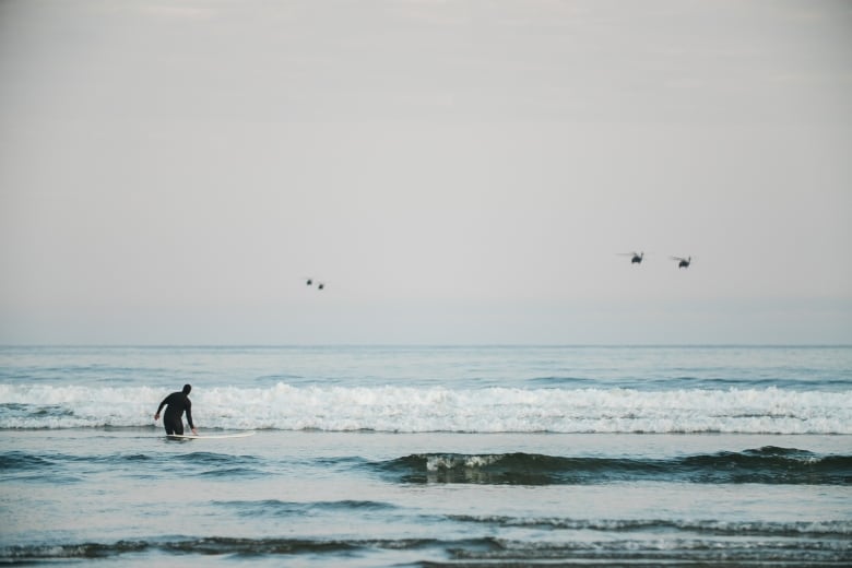 A surfer watches as four helicopters fly overhead of Long Beach near Tofino, B.C. on June 20, 2023.