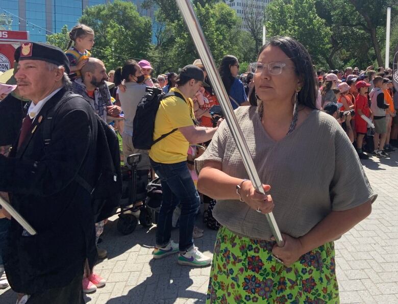 A woman in a grey shirt and green ribbon skirt carries a flag.