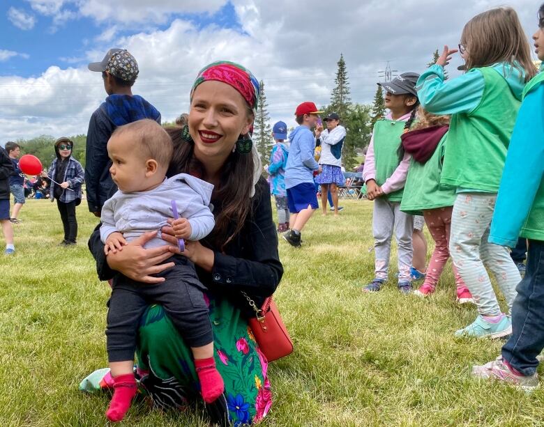 A woman in a ribbon skirt holds a toddler on her knee as she kneels in the grass.