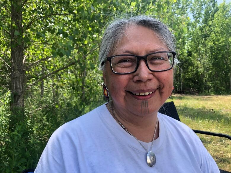 A Cree woman elder with four lines tattooed on her chin smiles into the camera beside a forest.