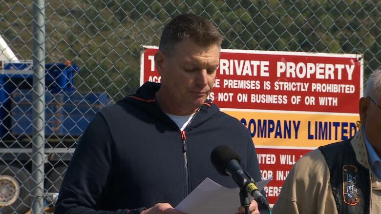 A man in a navy blue sweater looks down at his notes on paper while standing in front of a microphone.