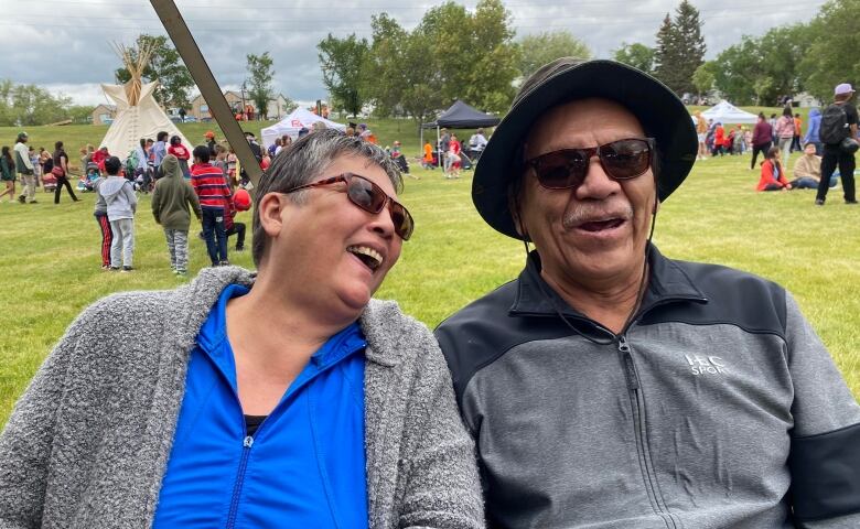 A woman and man, both dressed in grey, smile and laugh while they sit on a bench.