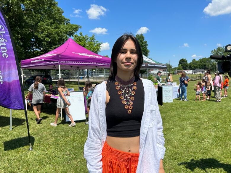 A 20 year old girls standing in a black top and white blouse outdoors. Event tents are behind her. 