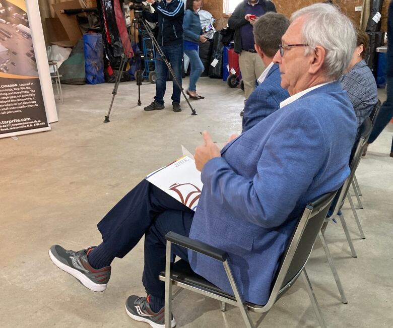 A man with short grey hair and glasses is seated in a chair at an event. He's wearing an indigo coloured suit jacket over a white shirt, dark dress pants and grey and white running shoes.