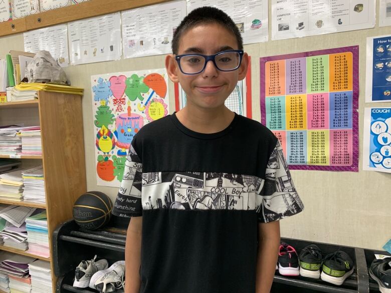 A little boy in glasses and a black and white shirt smiles at the camera in a classroom.