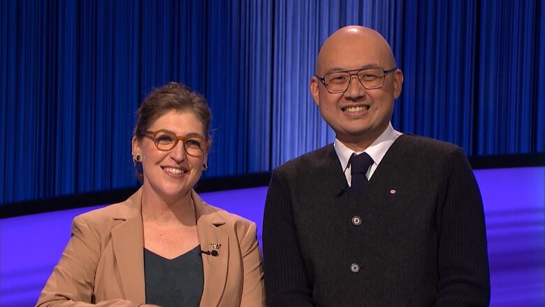 Two people stand side by side facing the camera in front of a blue backdrop