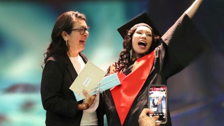 A girl in a graduation gown happily raises her hand in the air while accepting a piece of paper. 