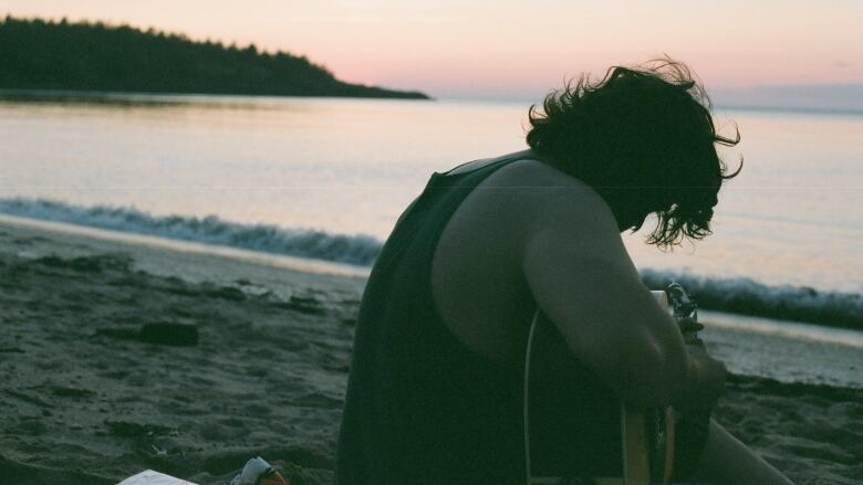 A man with an acoustic guitar sits on a beach as the sun sets.