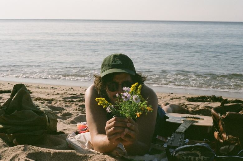 A mean wearing sunglasses and a hit sits on the beach and looks at the camera. He has some flowers in his hands.