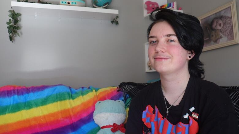 A teenage boy sits on a couch beside a rainbow coloured blanket.