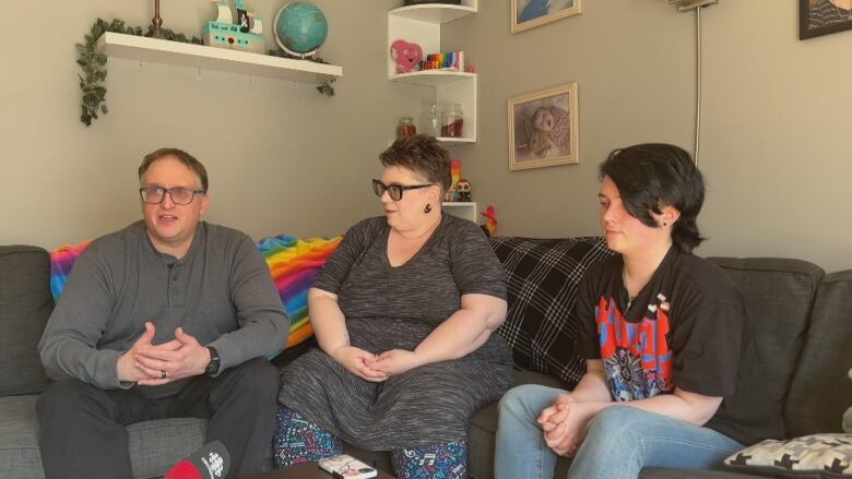 Two men and a woman sit on a couch with a wall behind them.