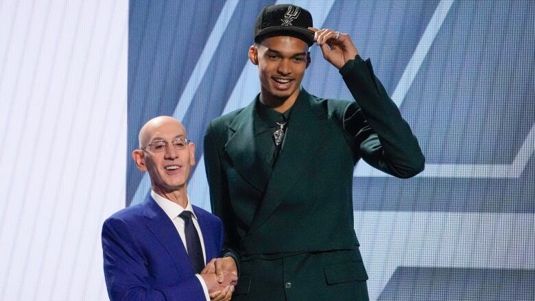 A male basketball player smiles while wearing a suit and team hat as he shakes right hands with a shorter man in a suit.