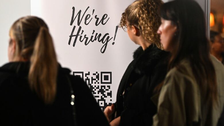 Women are shown at a job fair in front of a hiring sign.