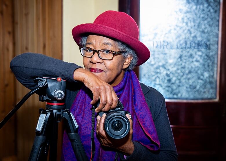 A Black woman leaning on a camera tri-pod with a red hat and purple scarf.