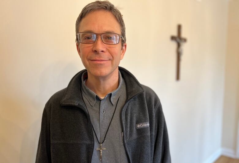 A man with greyish hair, wearing glasses and a grey shirt and jacket, stands in front of a crucifix on a wall.