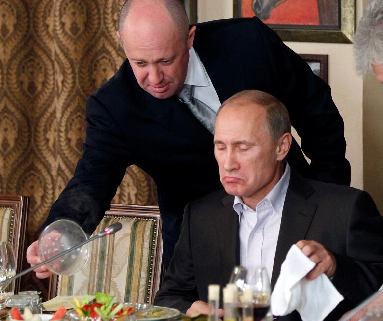 A man serves a dish of food to another man seated at a table.