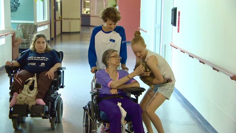 A girl pushes a woman in a wheelchair while another girl holds a cat.