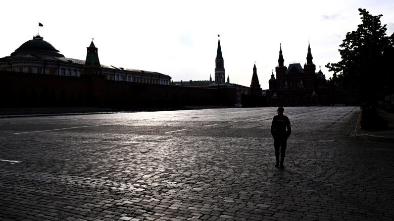 A sole person is seen in shadow, walking across the cobblestones of an expansive, empty public square, with buildings in the background.