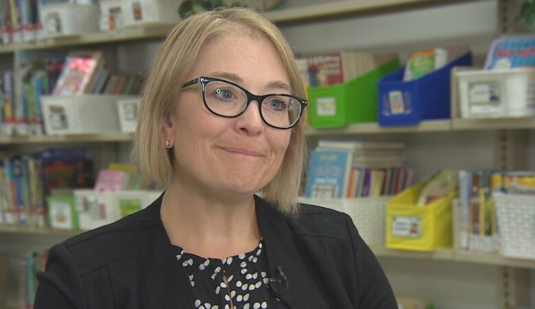 A woman with short blonde hair and black glasses is wearing a black and white top with a black blazer overtop. 