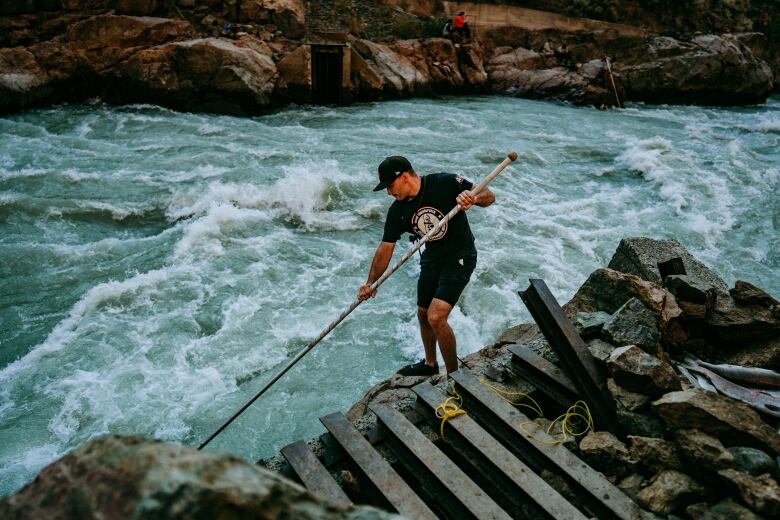 A man in black shorts and t-shirt fishes next to a river with a long spear. 