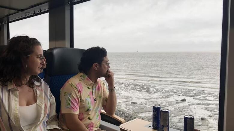 A woman and a man seated side by side on a train look out the window at a frothy white river.