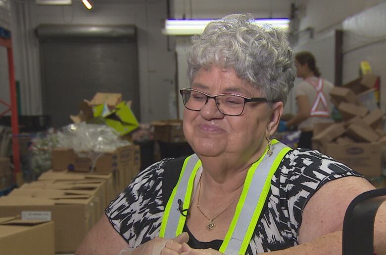  An older woman with short grey hair, glasses and wearing an apron is being interviewed in a warehouse. 