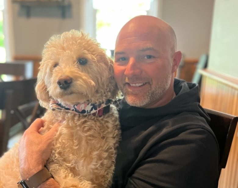 Robin Croucher smiles in a black hoodie with his dog Adley.