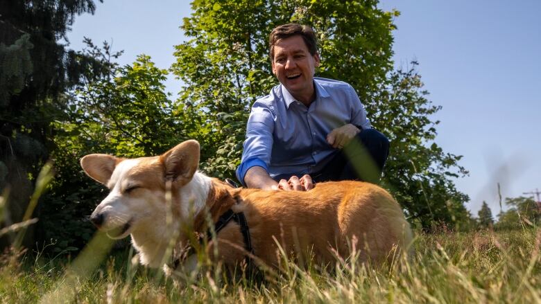 B.C. Premier David Eby wearing a blue dress shirt, smiles as he bends down to pet a white and blonde corgi dog named Honey.