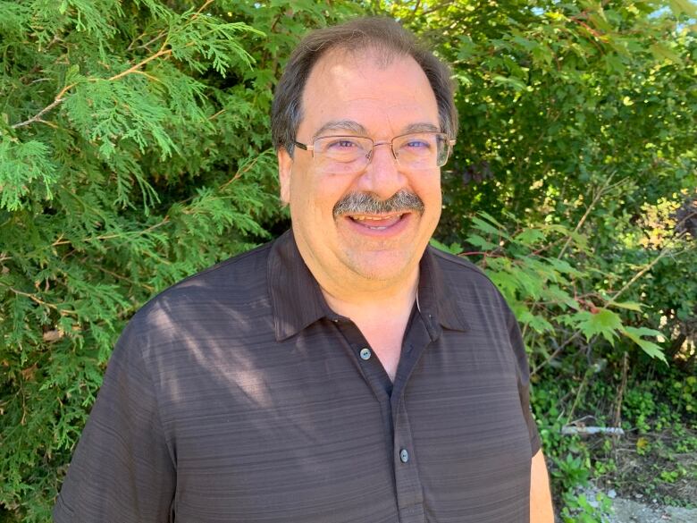 A man with a moustache and eyeglasses smiling at the camera. He's standing in front of a green bush. 