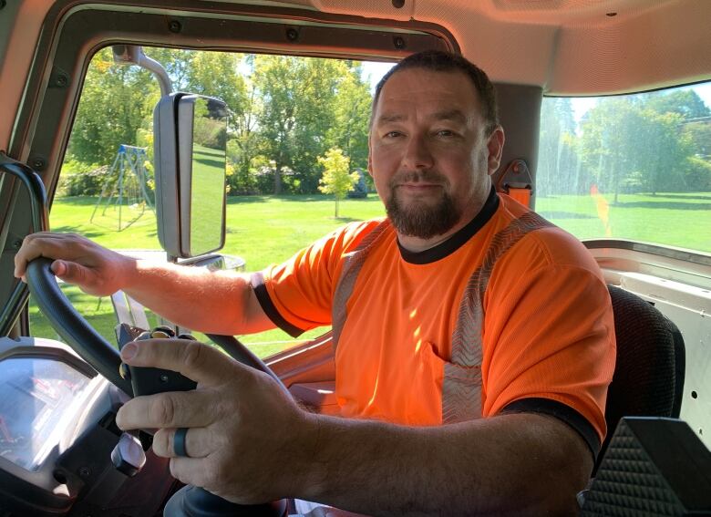 A man in an orange t-shirt behind the wheel of a huge truck, inside the cab. 