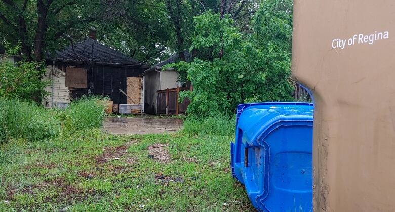 Garbage bins sit in front of the back lawn of a burned out house. The windows and door is placarded; the roof and back wall of the structure is charred. Shrubs are growing in the yard.